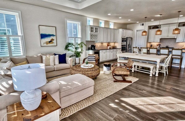 A spacious living room with a sofa, ottoman, and coffee table, a dining area with a table and chairs, and a kitchen with white cabinets and a refrigerator.