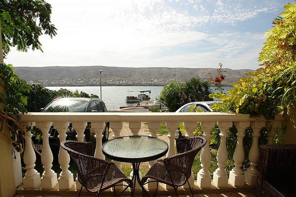 A terrace with a round table and two wicker chairs, overlooking a body of water and a distant cityscape.