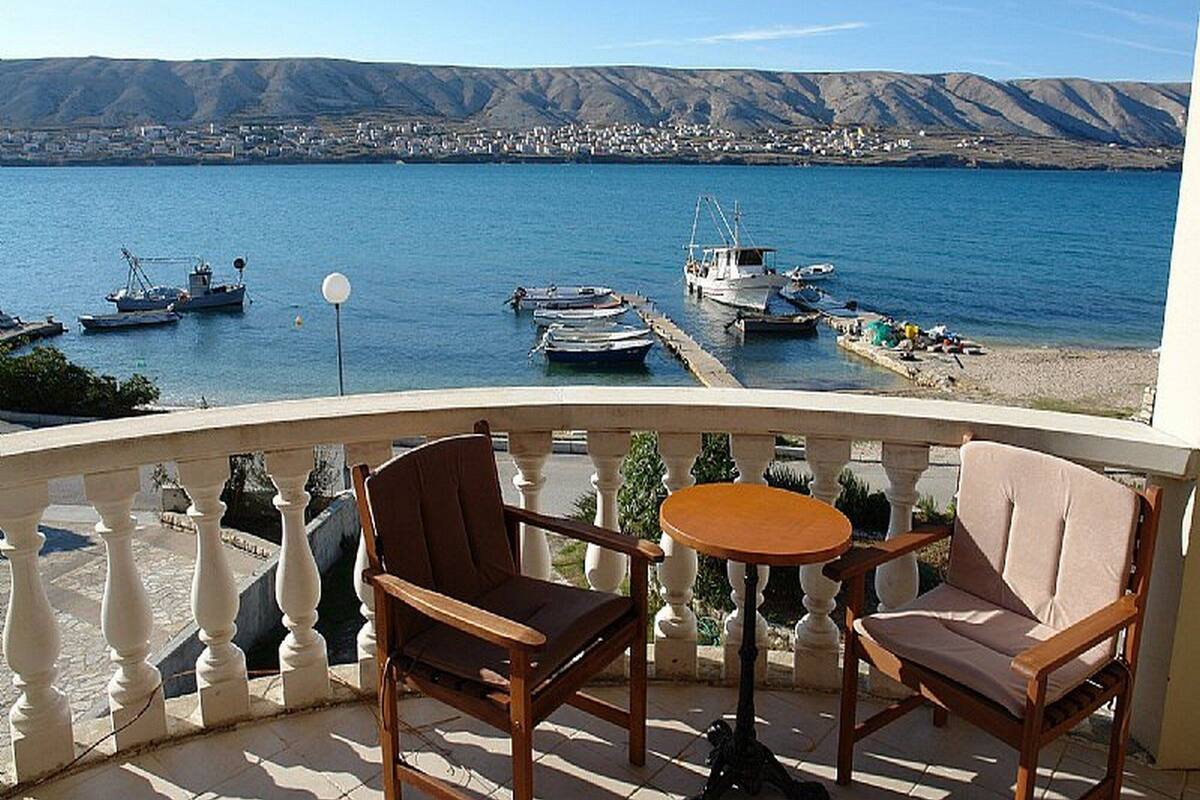 A balcony with two wooden chairs and a small round table, overlooking a marina with boats and a distant town.