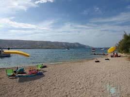 Una spiaggia con ciottoli, due ombrelloni gialli, una sedia verde e un asciugamano da spiaggia colorato. Si vedono imbarcazioni sull'acqua.