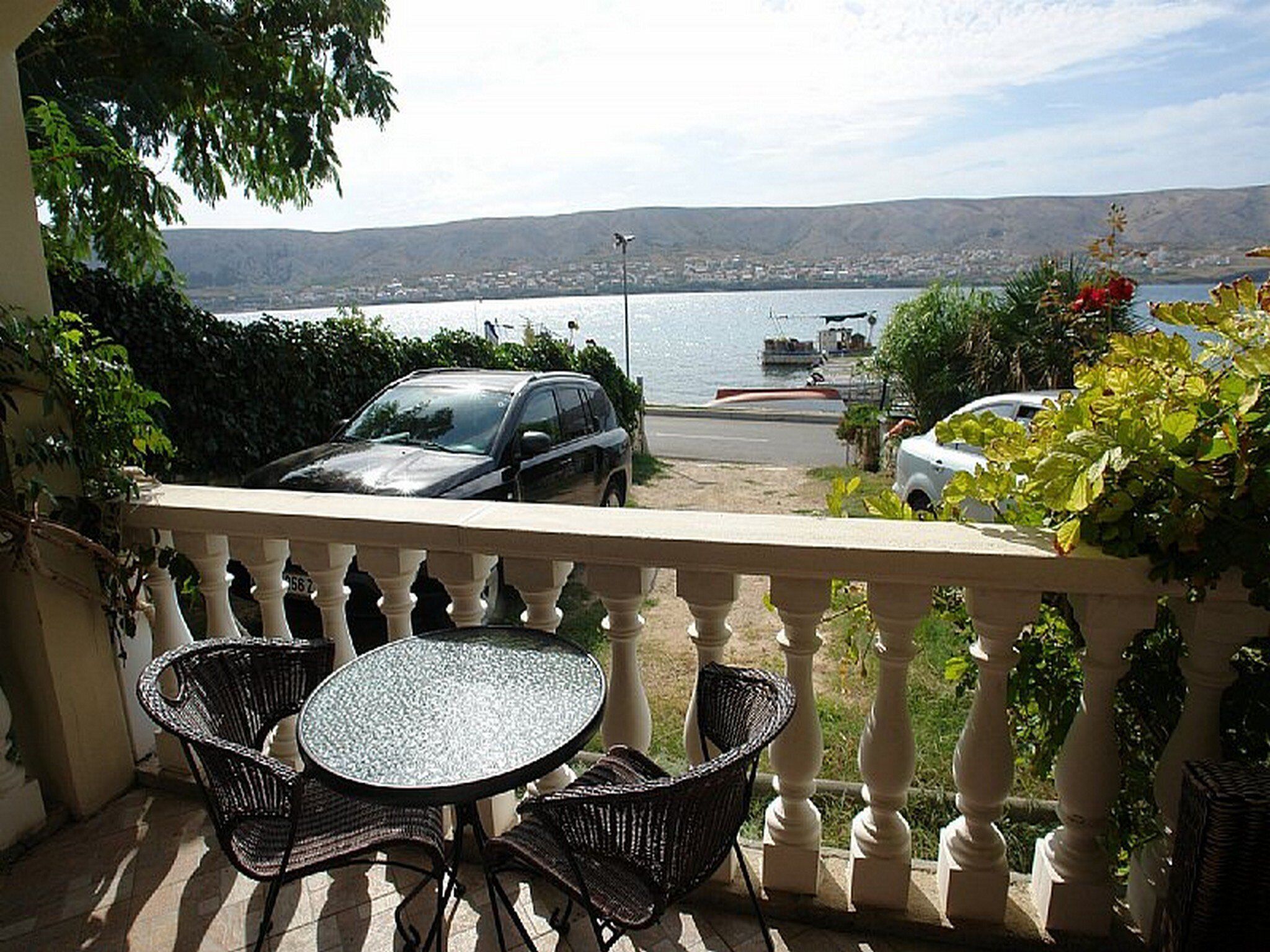 A balcony with a table and chairs, overlooking a body of water with boats and a distant cityscape.