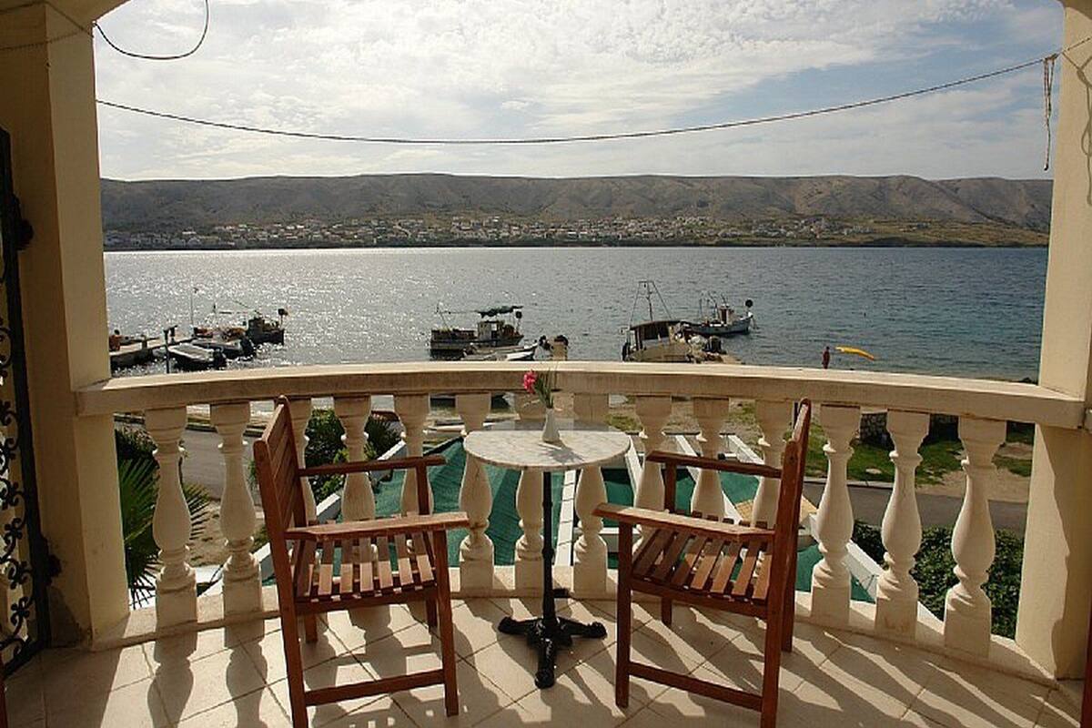 A balcony with two wooden chairs and a small round table, overlooking a calm lake with boats and a distant shoreline.