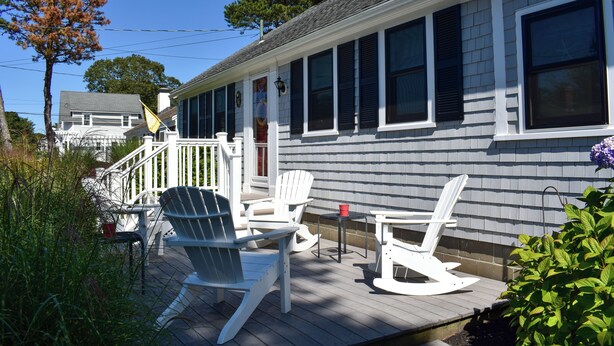 Um deck de madeira com cadeiras Adirondack brancas, uma mesa pequena e um balde vermelho.