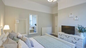 Un dormitorio con cama, mesita de noche, lámpara, cómoda, televisor y espejo.