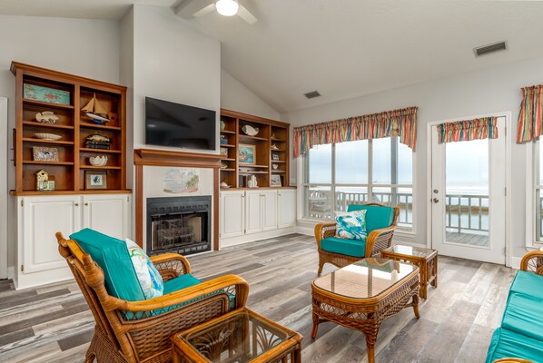 Amplia sala de estar con muebles de mimbre, chimenea y ventanas grandes con vista al océano.