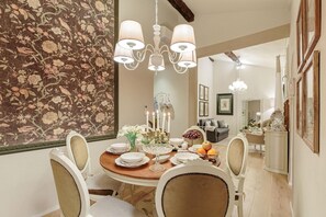 A dining area with a round table set for four, surrounded by white upholstered chairs. A chandelier hangs above the table, and a floral wallpaper is visible on the wall. - Casa Uccellini in Lucca (Lucca)
