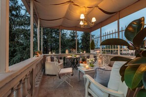 A covered outdoor seating area with wicker furniture, a table with a candle, and a view of trees and a fence. - Casa Uccellini in Lucca (Lucca)