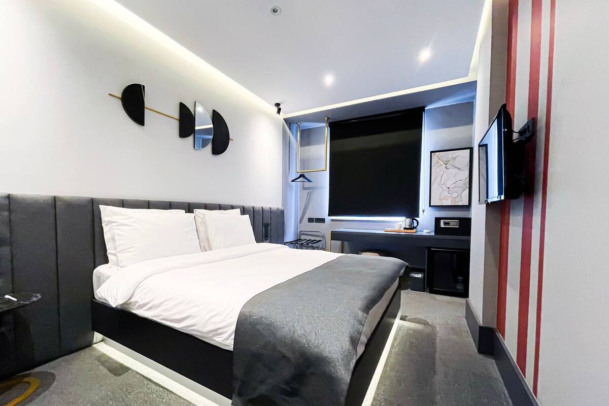 A modern hotel room with a large bed, a desk with a flat-screen TV, a desk lamp, and a wall-mounted TV.