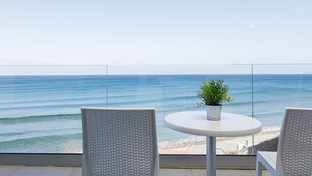 Un balcón con dos sillones blancos de mimbre y una pequeña mesa redonda con una planta en maceta, con vista a una playa y el océano.