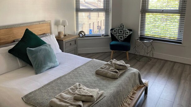 Une chambre à coucher comprenant un lit, une table de chevet, une chaise et deux fenêtres équipées de stores.