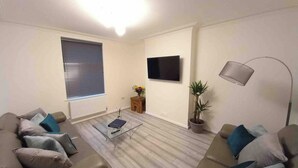 Un salon comprenant un ensemble de canapés, une table basse, un téléviseur fixé au mur, une lampe sur pied et une plante en pot.