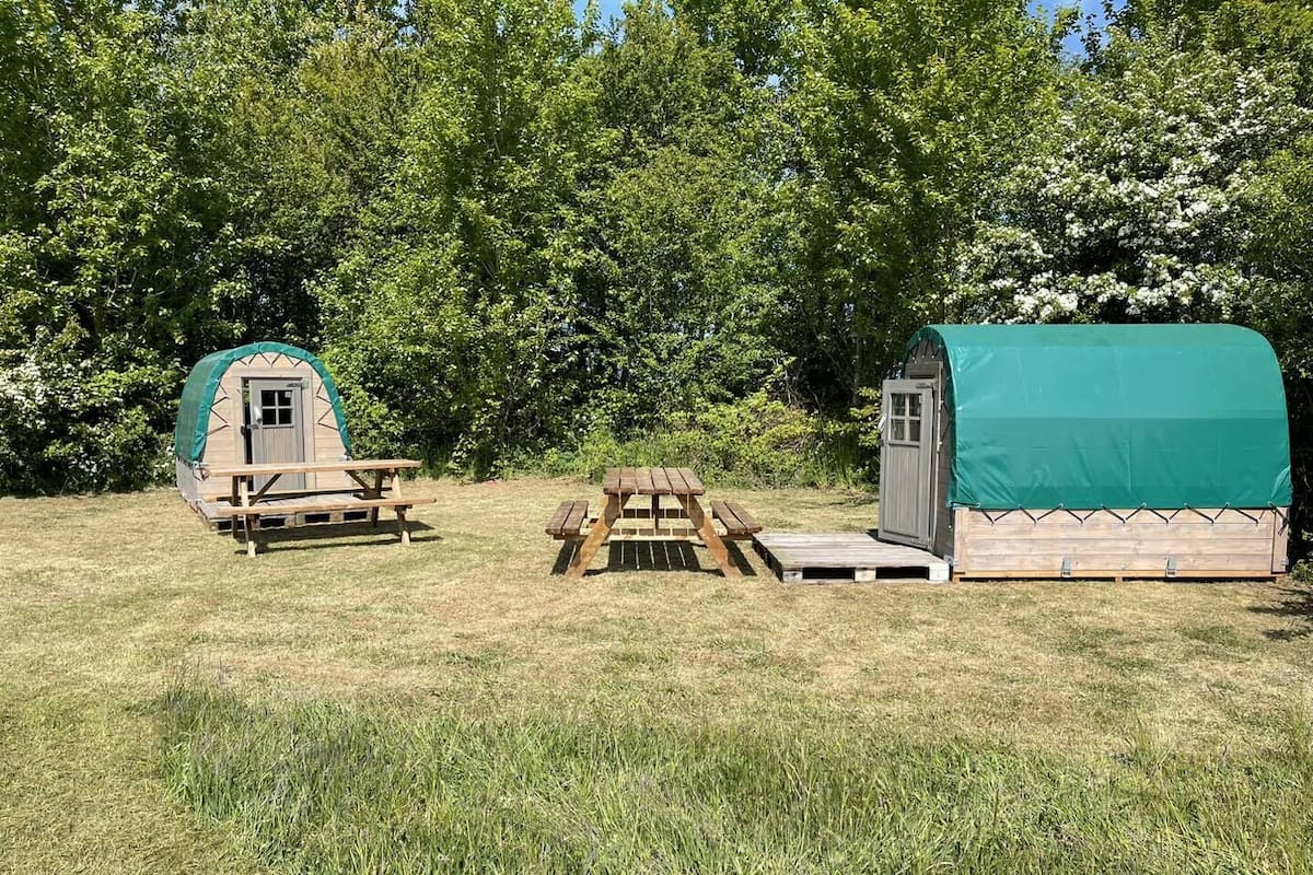 A small, rustic wooden structure with a green roof, a wooden bench, and a picnic table in a grassy field.
