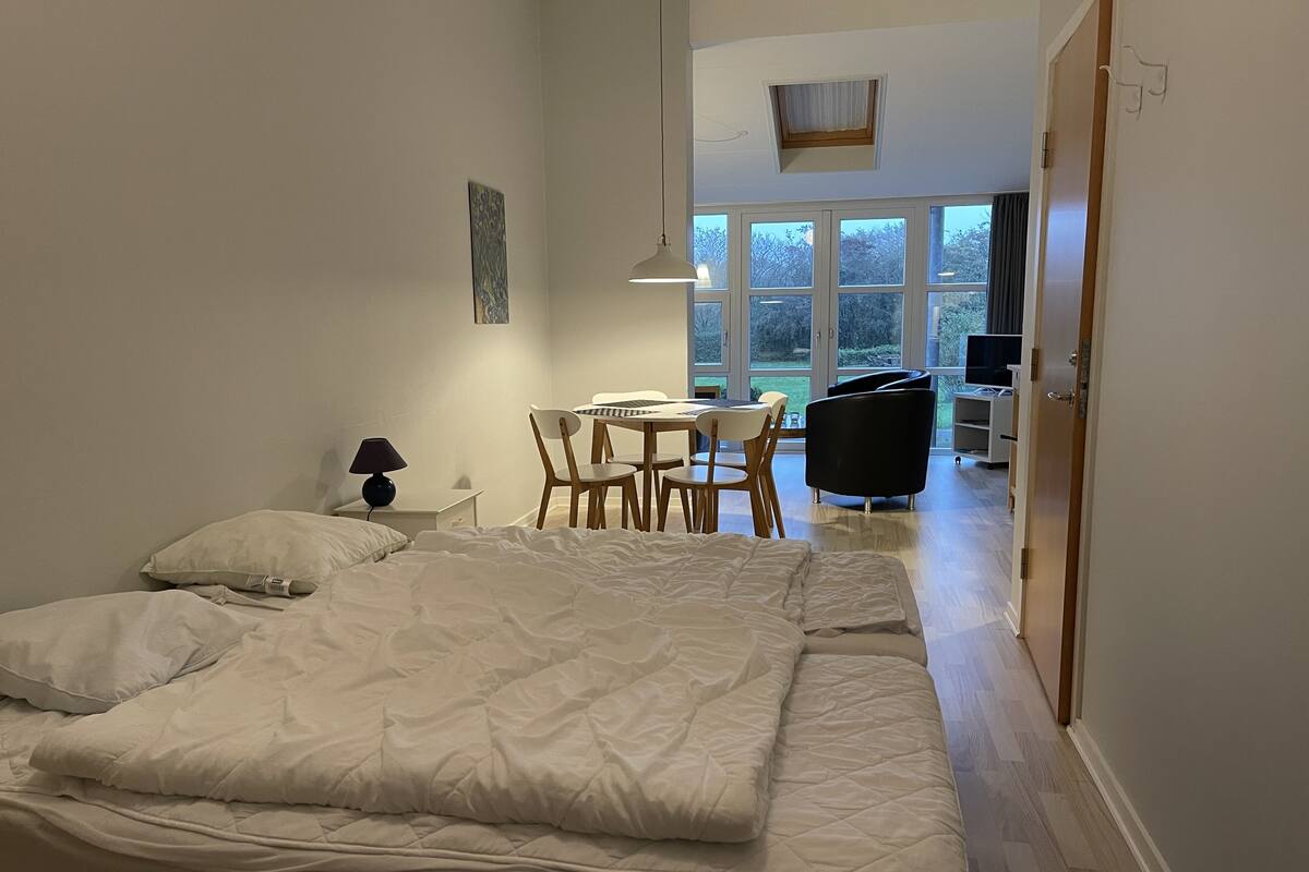 A bedroom with a bed, a desk, and a chair. There is a window with a view of trees and a door leading to another room.