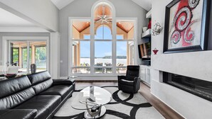 Un salon spacieux avec un canapé en cuir noir, une table basse en verre et une cheminée. Une grande fenêtre offre une vue sur l’extérieur.