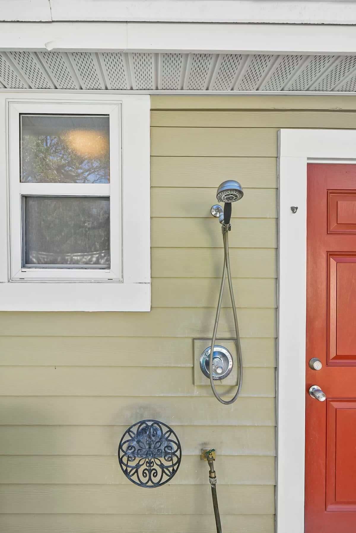 A house with a red door, a window with a view of trees, a shower with a hose, and a decorative plate.