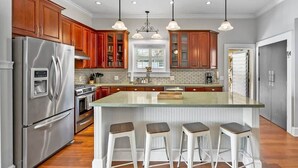 A modern kitchen with stainless steel appliances, wooden cabinets, a large island with bar stools, and pendant lighting. - Beach House, Pool, Golf Cart, Game Room, BBQ, (St. Augustine)
