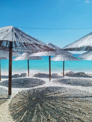 Una playa con varias sombrillas de paja y una orilla pedregosa.