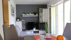 A modern living room with a sofa, a dining table with fruit, a TV, and a bookshelf. - Apartment Marina (Punat)
