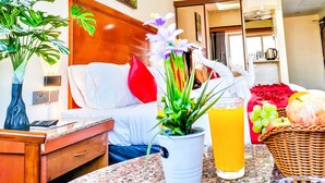 A well-lit room with a bed, a bedside table with a lamp, a vase with flowers, a fruit basket, and a glass of orange juice. - Le Paradise Palace Hotel Deira Dubai Al Rigga (Dubai)