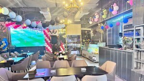 A restaurant with a large screen displaying a sports event, a bar area with a refrigerated display, and a seating area with tables and chairs. - Le Paradise Palace Hotel Deira Dubai Al Rigga (Dubai)