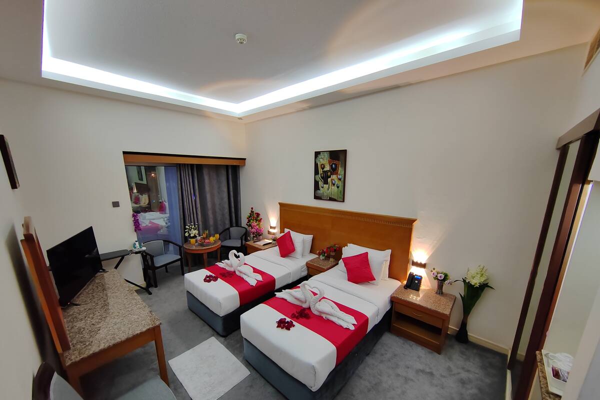 A hotel room with two beds, a desk, a chair, a small table with flowers, and a painting on the wall.