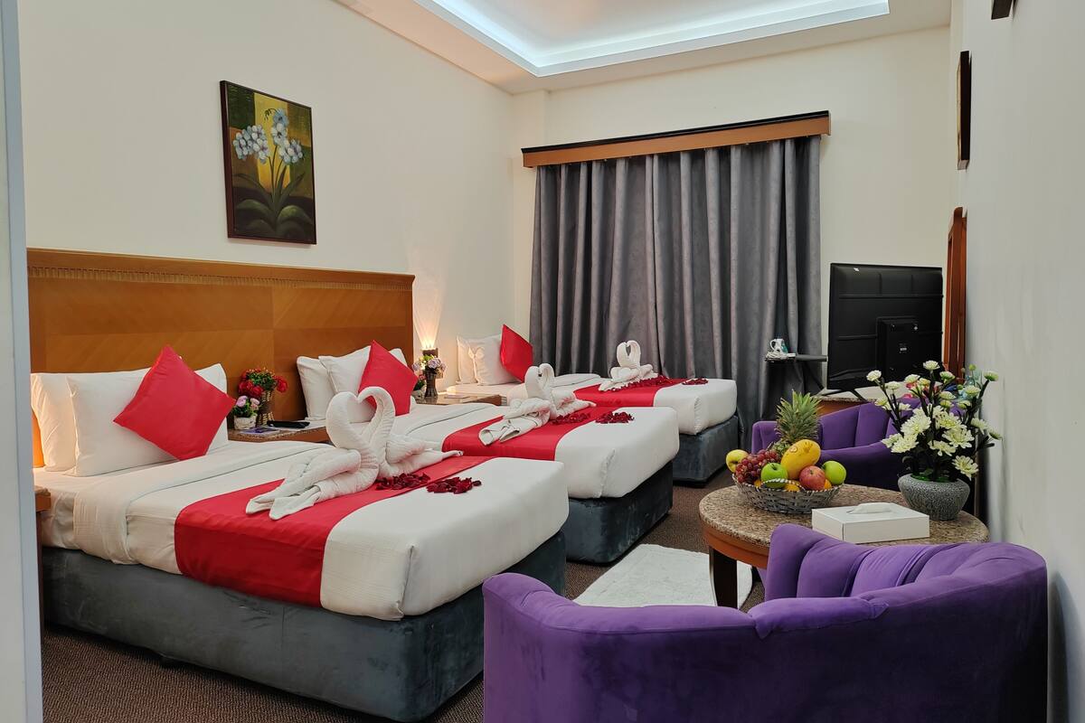 A hotel room with two beds, a TV, a small table with fruit, and purple chairs.