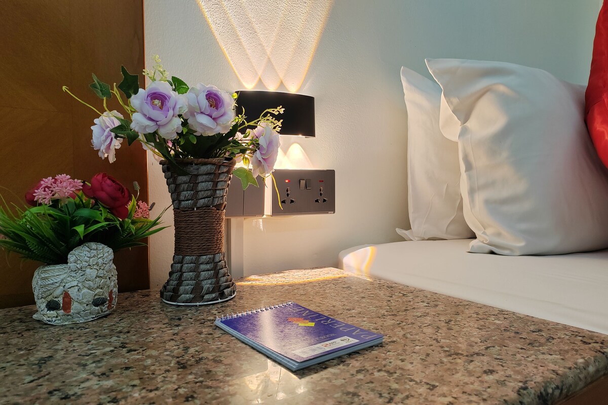 A neatly arranged room with a bed, a bedside table with a notebook, a vase with flowers, and a wall-mounted light.