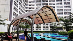 Zona de piscina con sillones de descanso y un área de estar techada, rodeada por un edificio de gran altura.