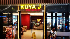 La entrada al restaurante Kuya J, decorada con adornos navideños, con un letrero de precaución en el suelo y una persona sentada en un banco afuera.