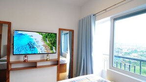 Una habitación de hotel con una cama, un televisor montado en la pared, una ventana con vista a la ciudad y un estante de madera con un farol.