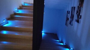 Een trap met blauwe verlichting, die leidt naar een plateau met een zwart gordijn en foto's in lijsten aan de muur.