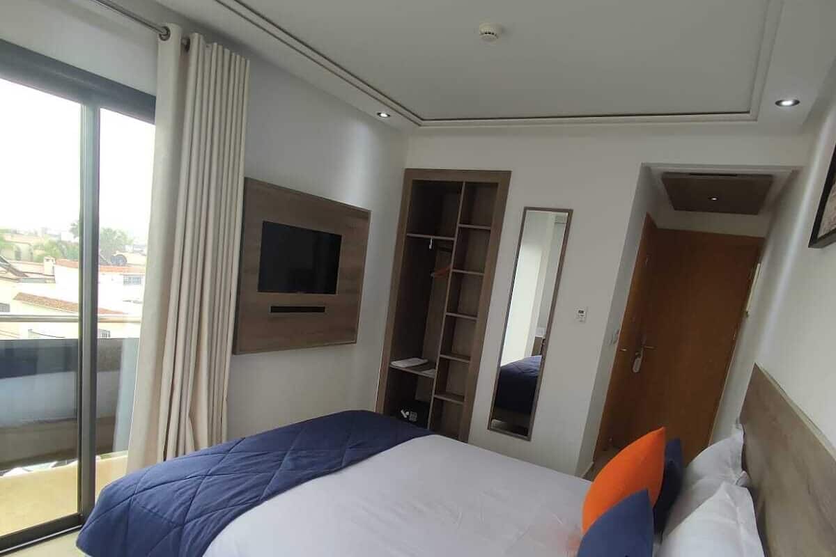 Habitación de hotel con una cama grande, televisor montado en la pared, armario empotrado y puerta corrediza de cristal al balcón.