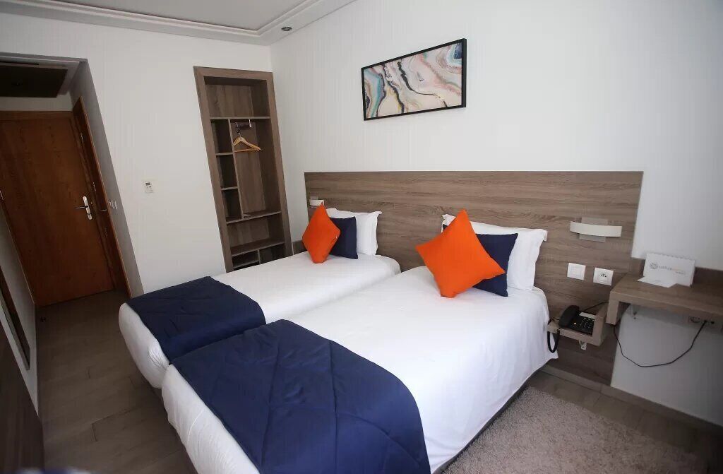 Quarto de hotel com duas camas, cabeceira de madeira, mesa de cabeceira com telefone e obra de arte na parede.