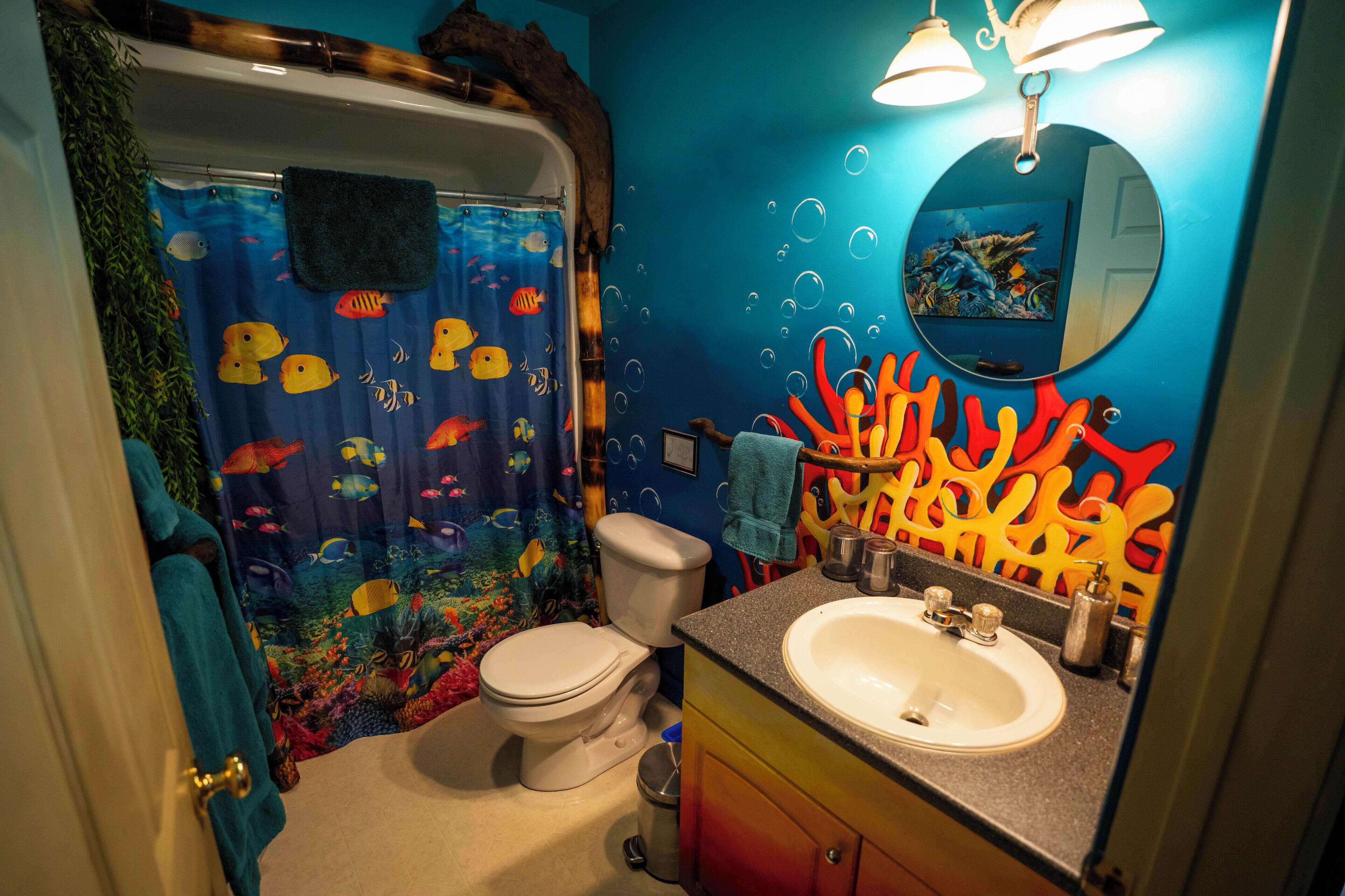 Une salle de bain avec un rideau de douche aux motifs colorés de poissons, des toilettes blanches, un lavabo avec un miroir et une décoration murale vive aux couleurs corail.