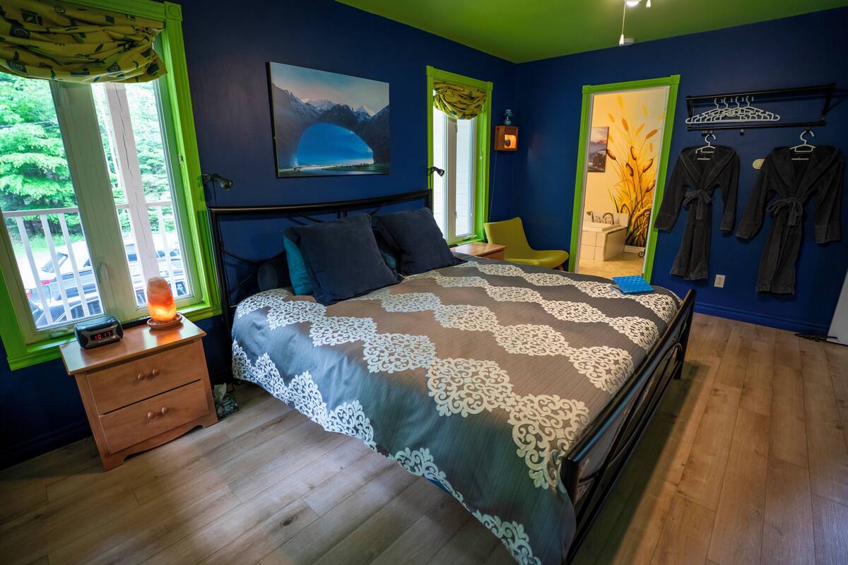 A bedroom with a bed, a nightstand, a window with curtains, a painting on the wall, and a rack with robes.