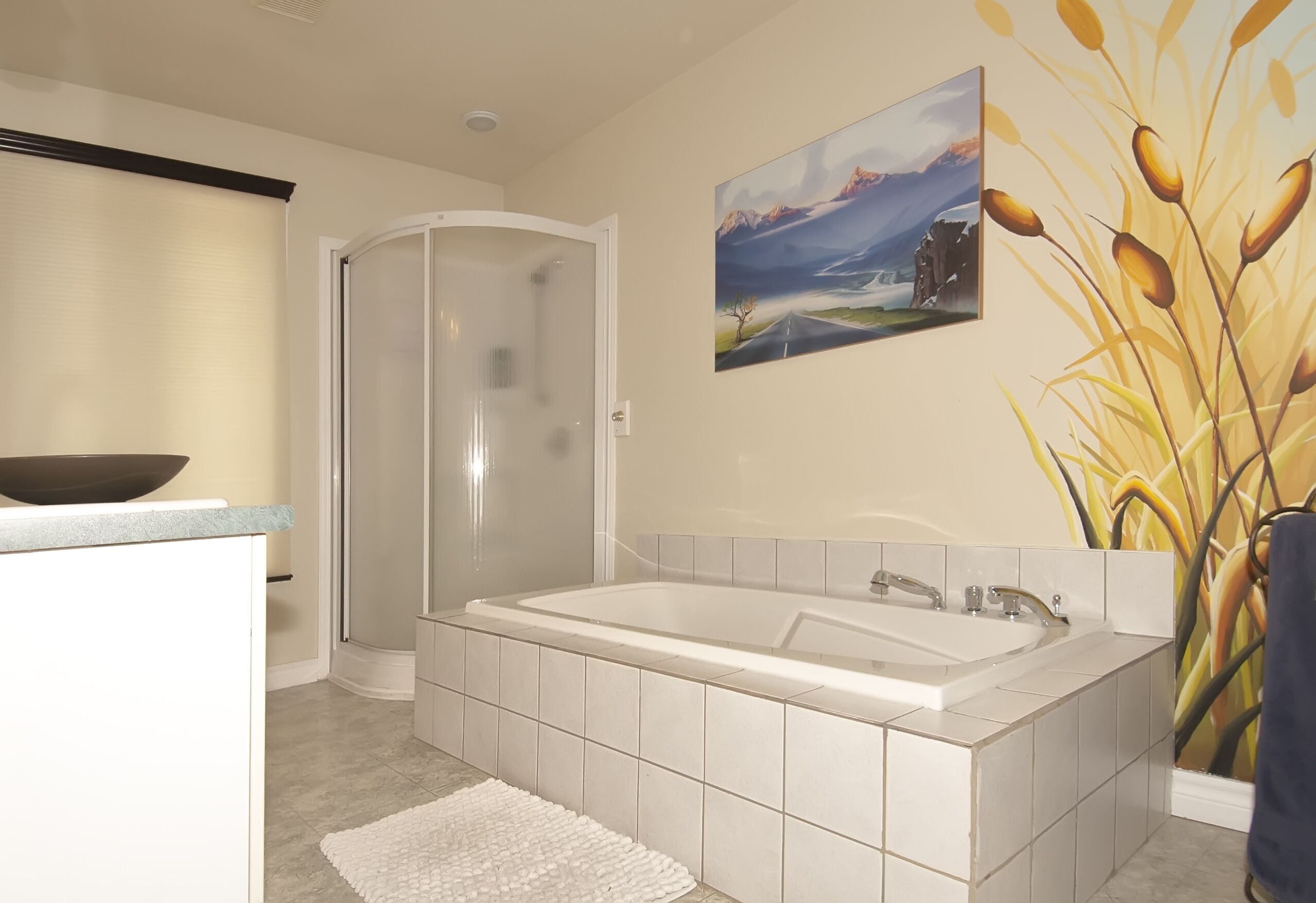 A modern bathroom with a large bathtub, a glass shower enclosure, and a painting of a landscape on the wall.