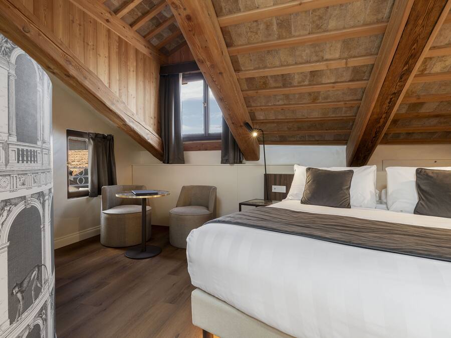 Un dormitorio con una cama grande, una mesita redonda pequeña, una silla y una ventana con cortinas.
