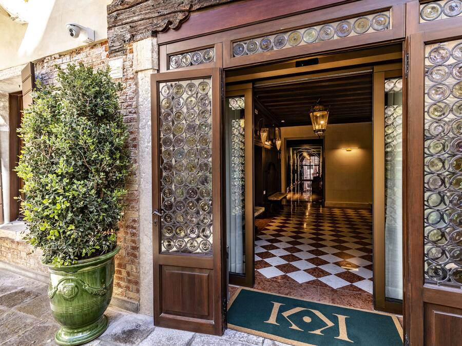 A hotel entrance with a checkered floor, a green doormat with a logo, and a potted plant.