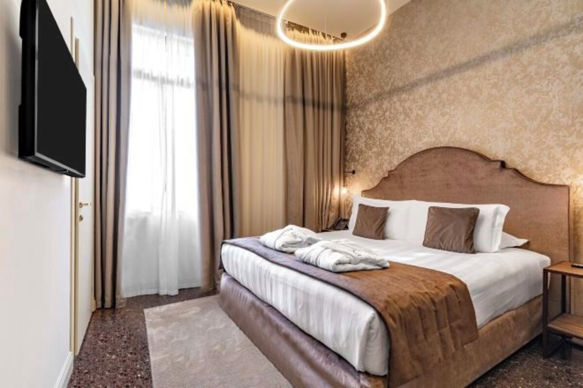 A hotel room with a large bed, a flat-screen TV mounted on the wall, a round light fixture above the bed, and a view of the outdoors through the window.