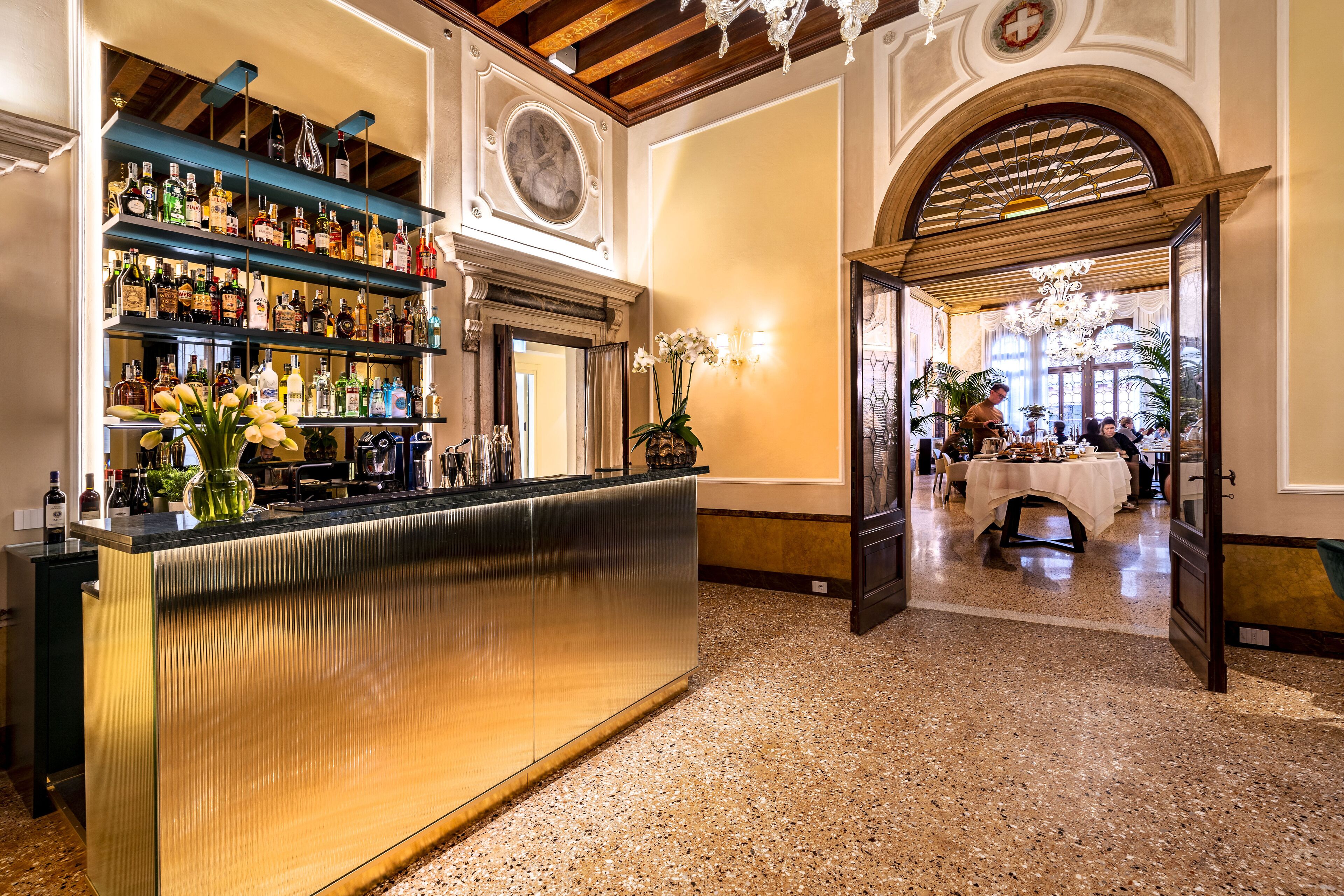 A bar area with shelves of liquor bottles, a polished counter, and a chandelier.