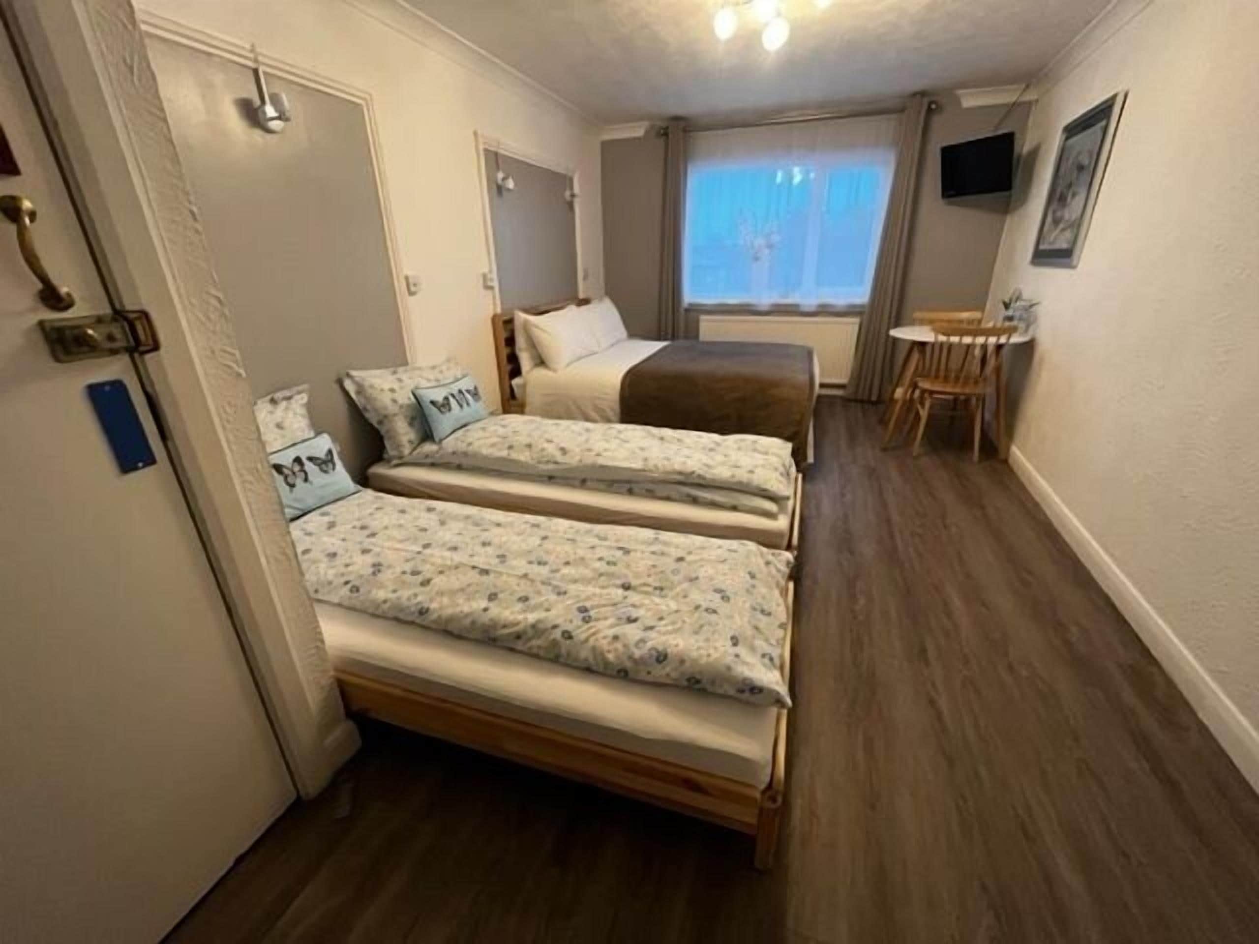 Una habitación con dos camas, una mesita, una silla y un ventanal con cortinas.