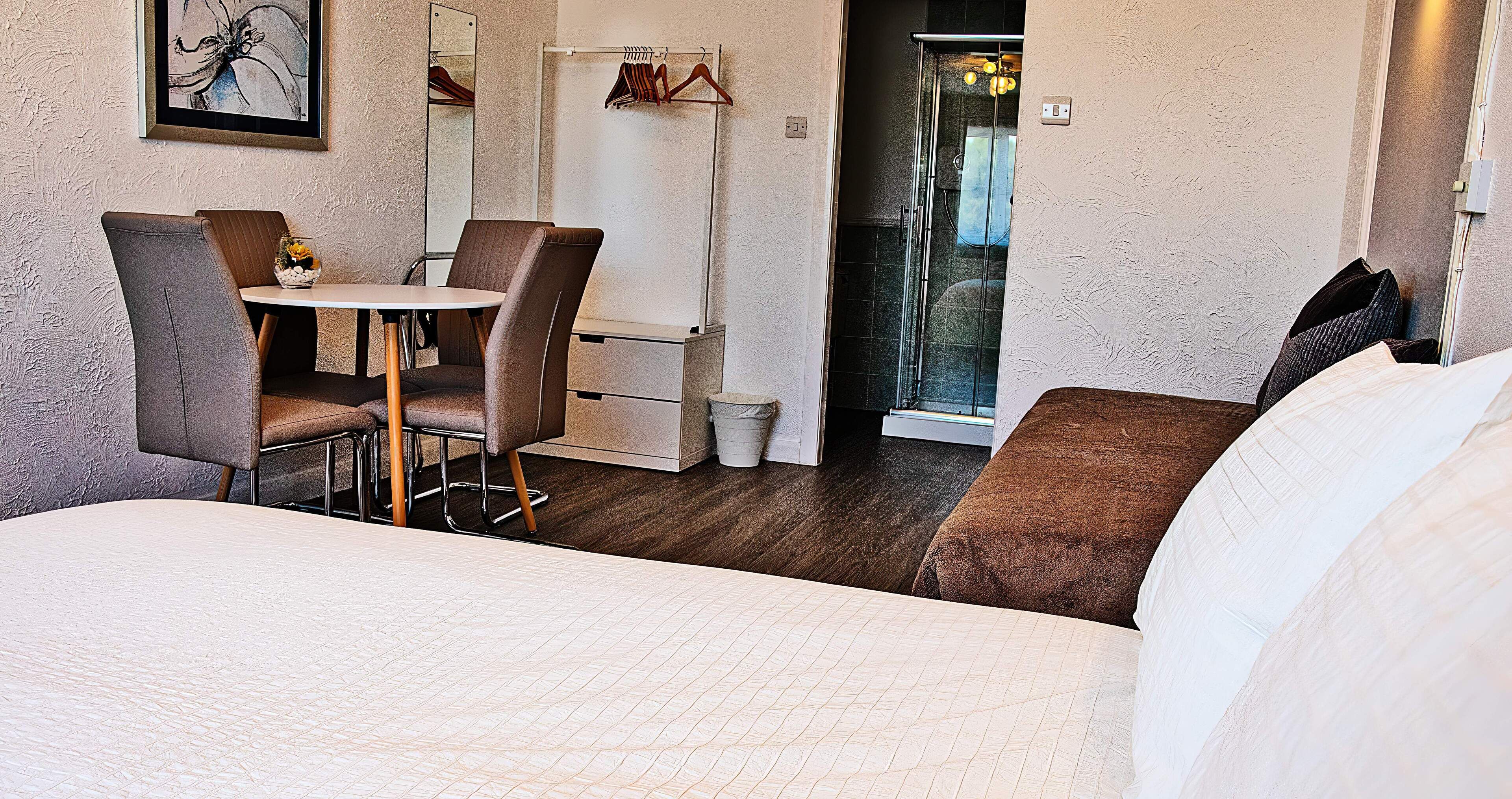 A hotel room with a bed, a dining table with chairs, a small table with a vase, a coat rack, and a shower area.