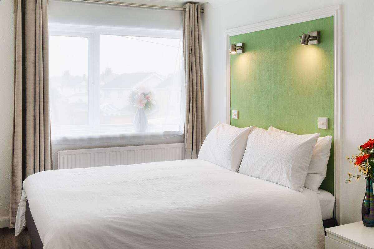 A neatly made bed with white linens in a room with a green accent wall, a window with curtains, and a vase with flowers on the nightstand.
