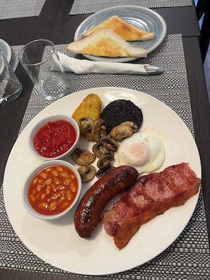 A plate of breakfast food including sausages, bacon, mushrooms, black pudding, toast, beans, and tomato sauce.