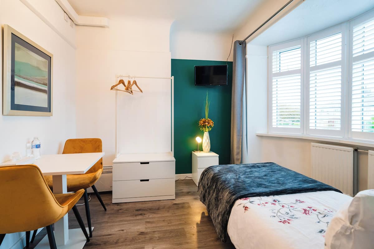 A modern bedroom with a bed, a desk with chairs, a TV, and a window with blinds.