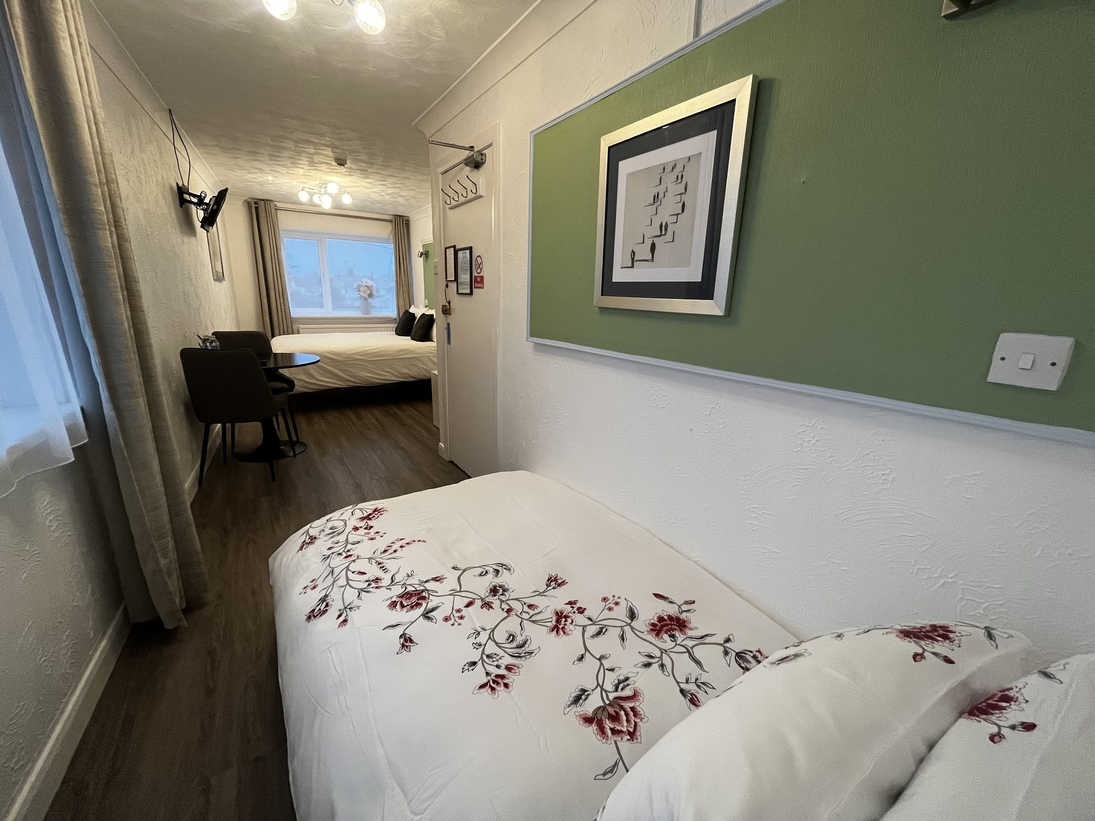 Une chambre d’hôtel avec un lit, un bureau et une chaise. Il y a une fenêtre avec des rideaux, un tableau encadré au mur et un interrupteur.