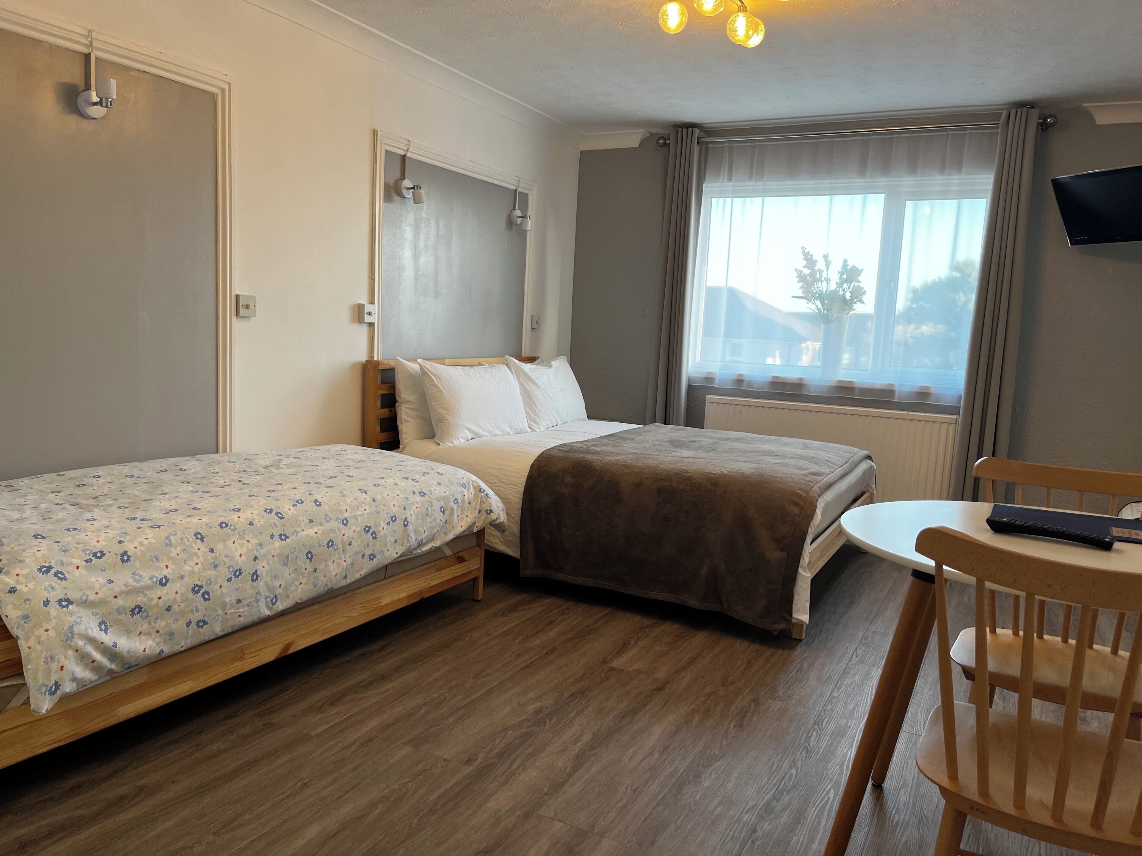 A hotel room with two single beds, a wooden chair, a small table, a TV on the wall, and a window with curtains.