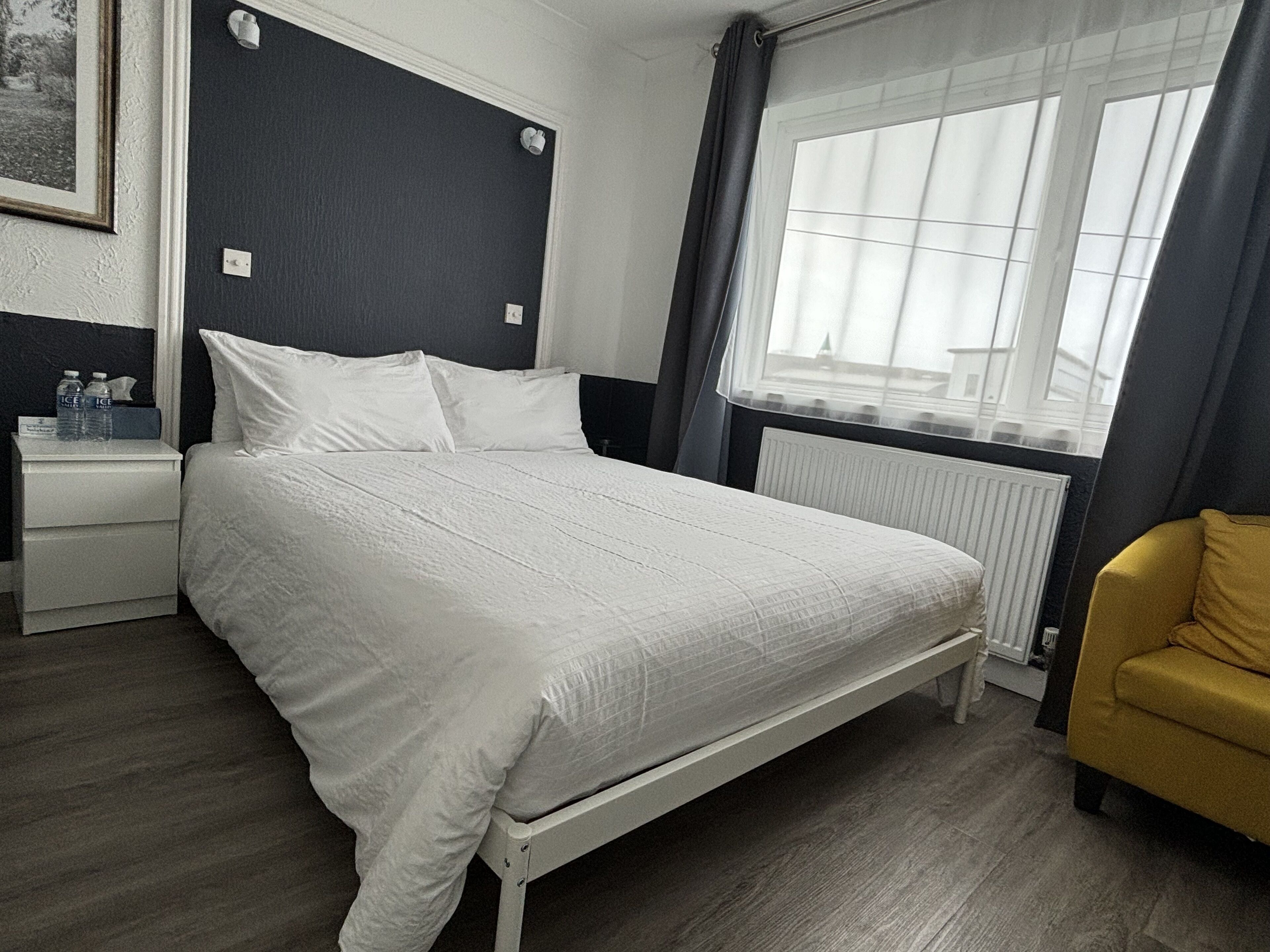 Een slaapkamer met een bed, een nachtkastje met waterflesjes, een radiator, een raam met gordijnen en een gele fauteuil.