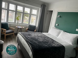 A hotel room with a bed, two armchairs, a small table, and a window with blinds.