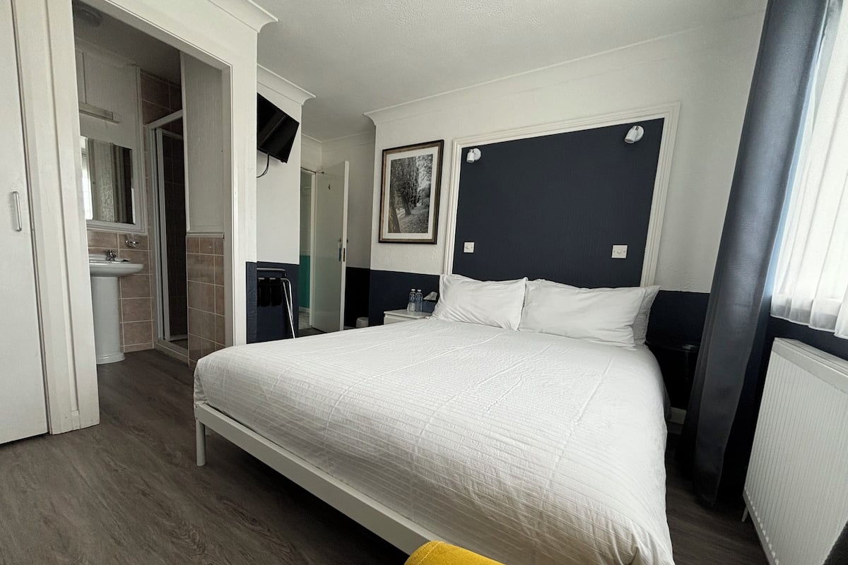 A hotel room with a large bed, a bedside table, a nightstand, a window with curtains, and a bathroom visible through an open door.